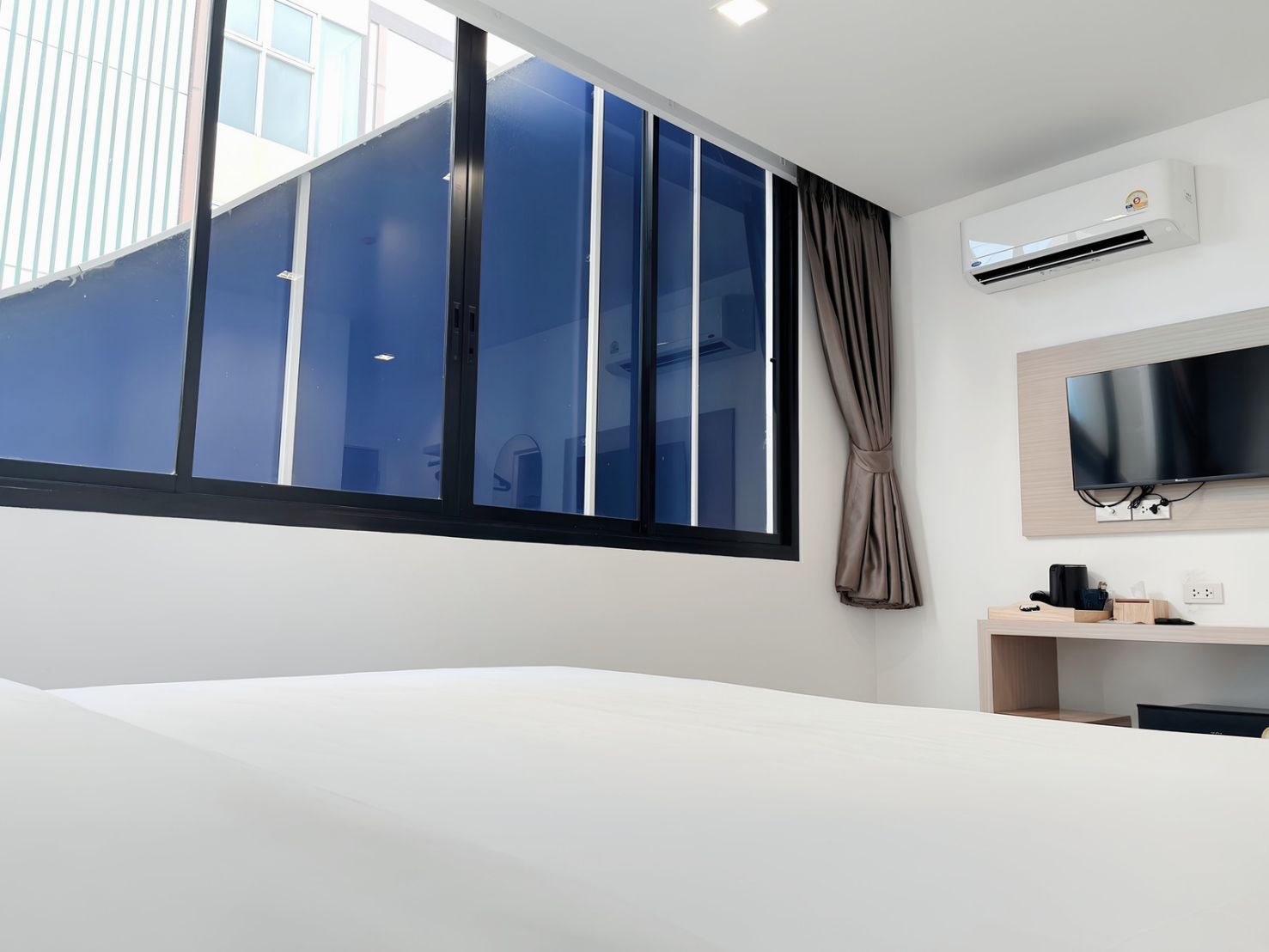 A hotel room with a large window, a flat-screen TV, a desk with a chair, and an air conditioning unit.