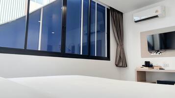 A hotel room with a large window, a flat-screen TV, a desk with a chair, and an air conditioning unit.