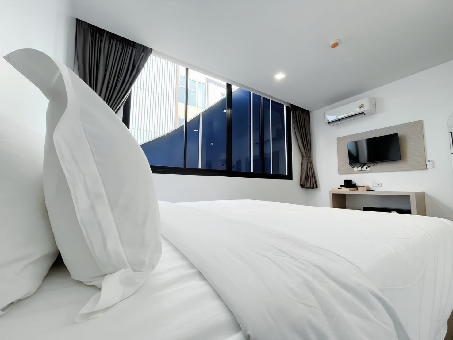 A modern hotel room with a large bed, a flat-screen TV, an air conditioning unit, and a window with curtains.