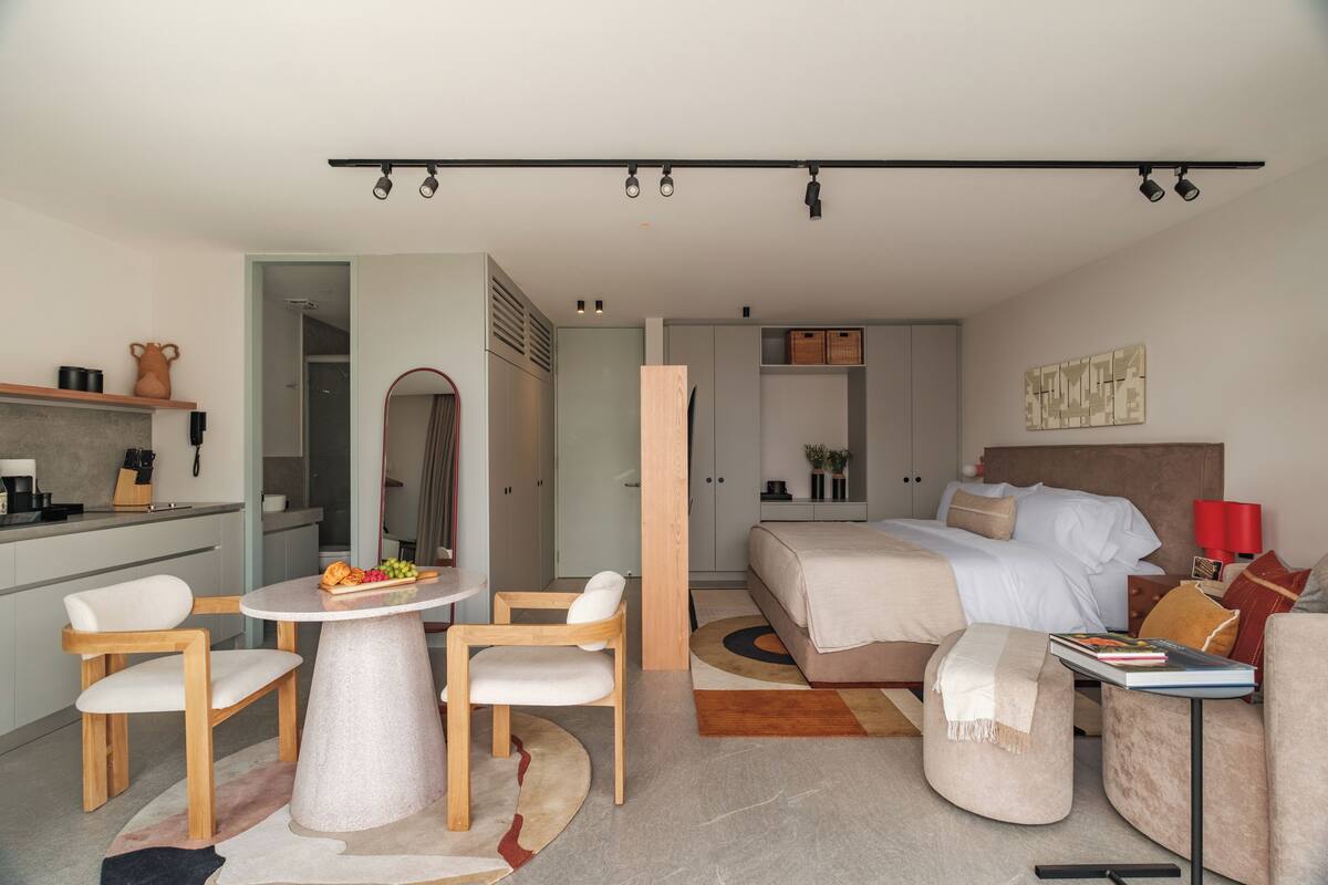 A modern hotel room with a bed, a dining area with chairs and a table, a kitchen area, and a bathroom.