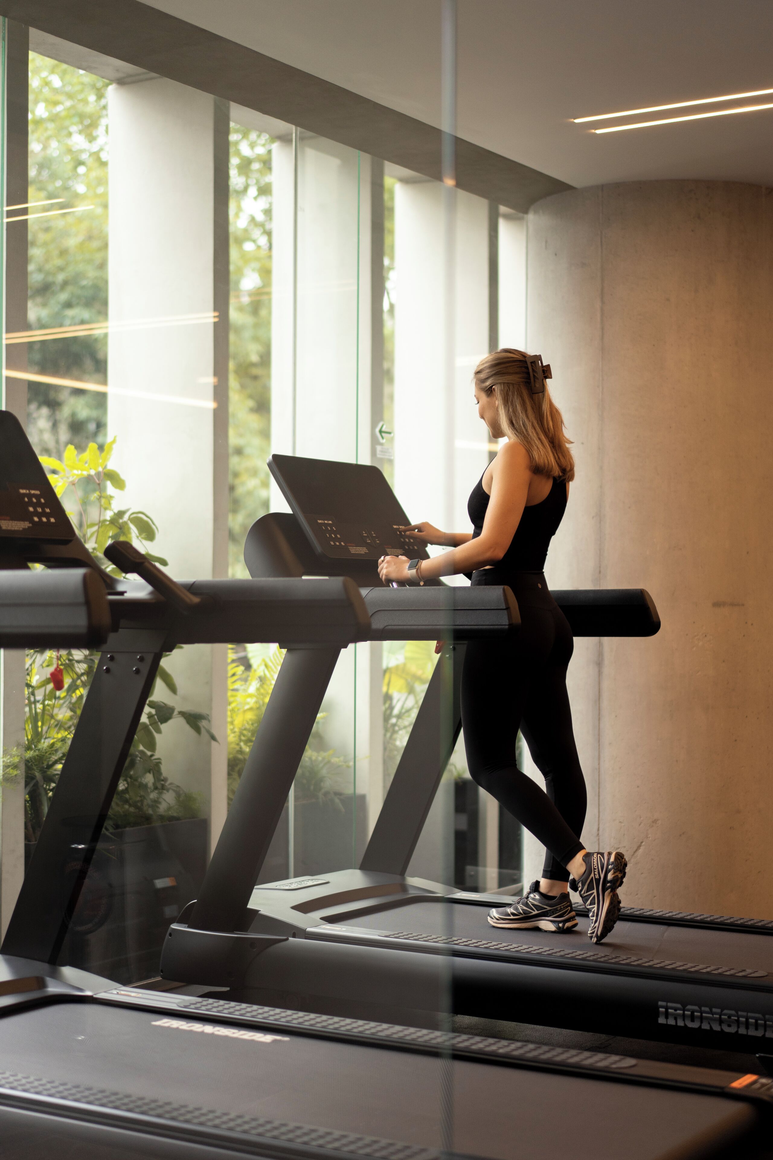 A woman using a treadmill in a modern gym with large windows and indoor plants.