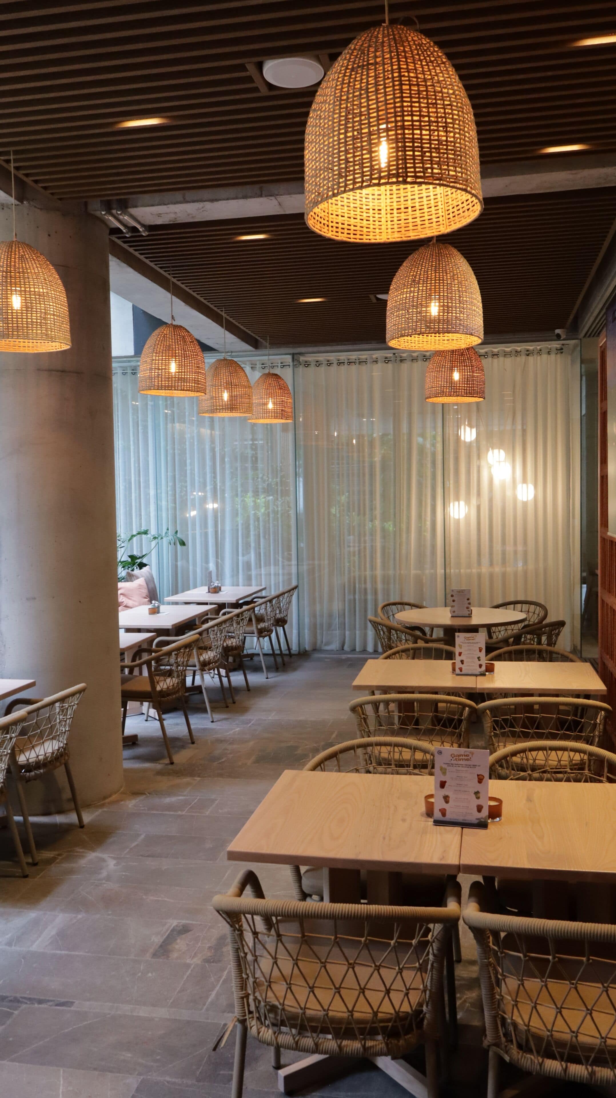 Ein Restaurantinnenraum mit Korbstühlen, Holztischen und gewebebespannten Pendelleuchten.