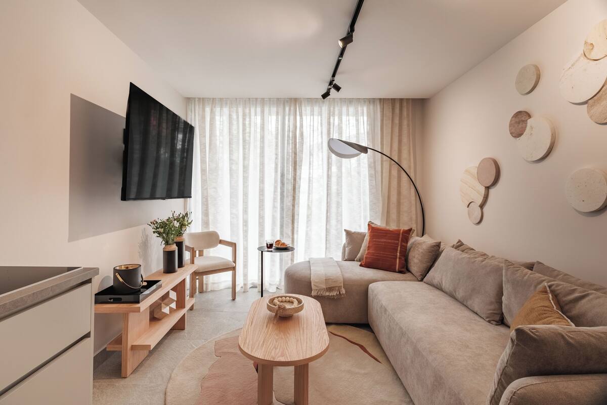 A modern living room with a sofa, a coffee table, a TV mounted on the wall, and a window with curtains.