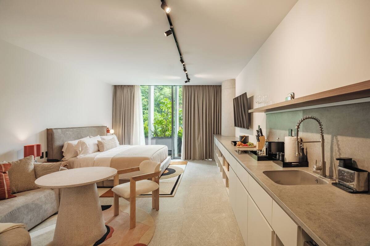 A modern hotel room with a bed, sofa, dining table, kitchen area, and a large window with curtains.