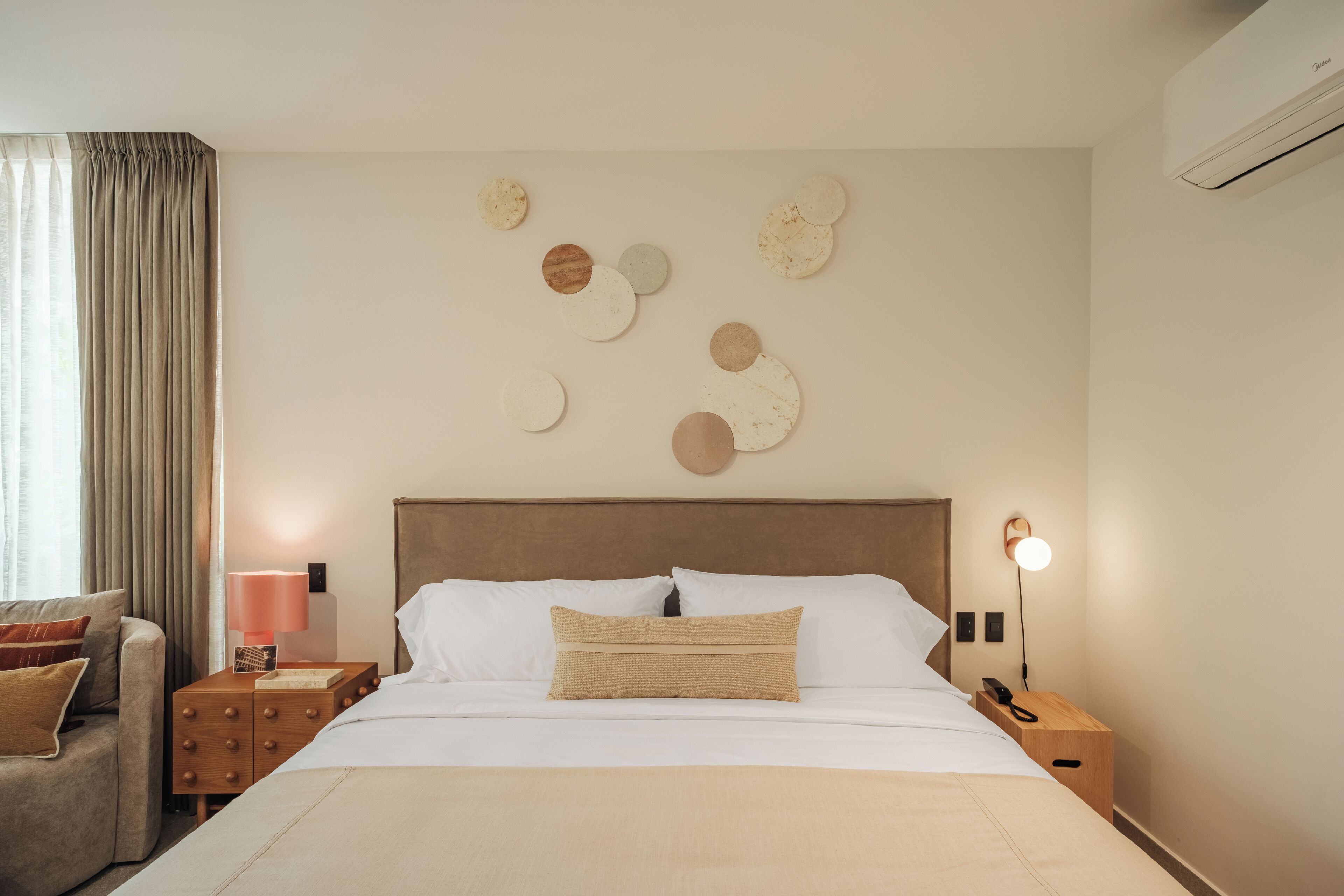 Quarto de hotel com cama, mesa de cabeceira, luminária, cadeira e parede com detalhes circulares.