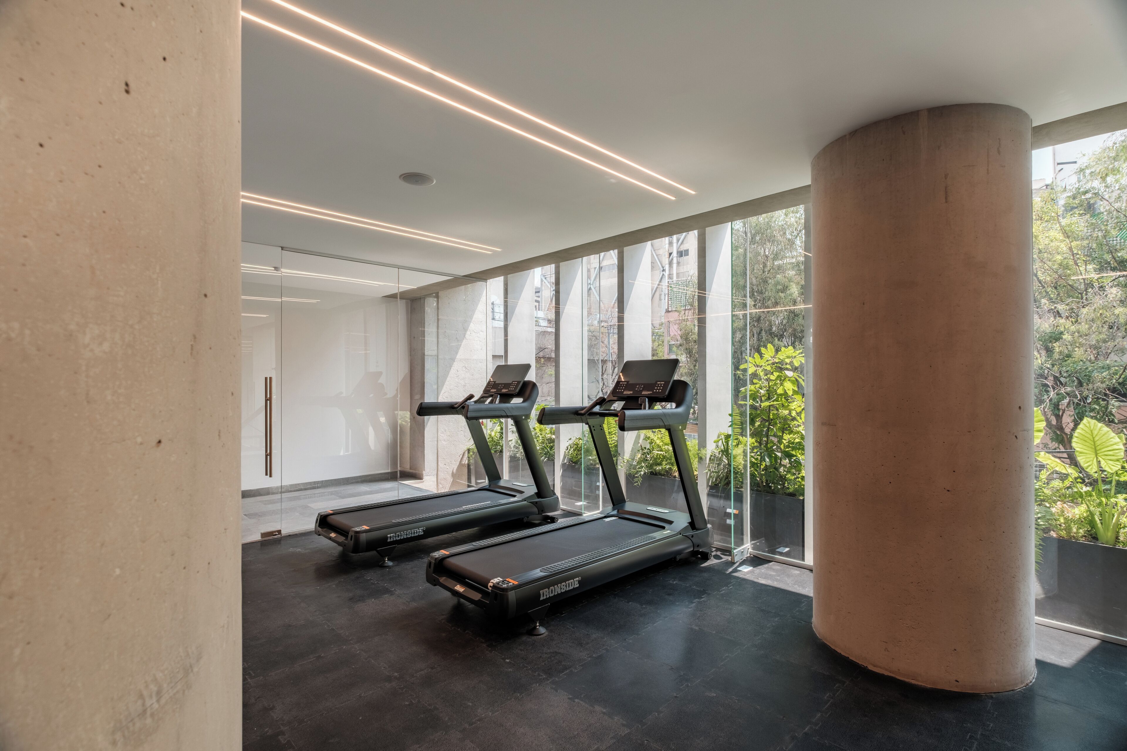 Een sportschool met loopbanden, grote ramen en een betonnen pilaar.