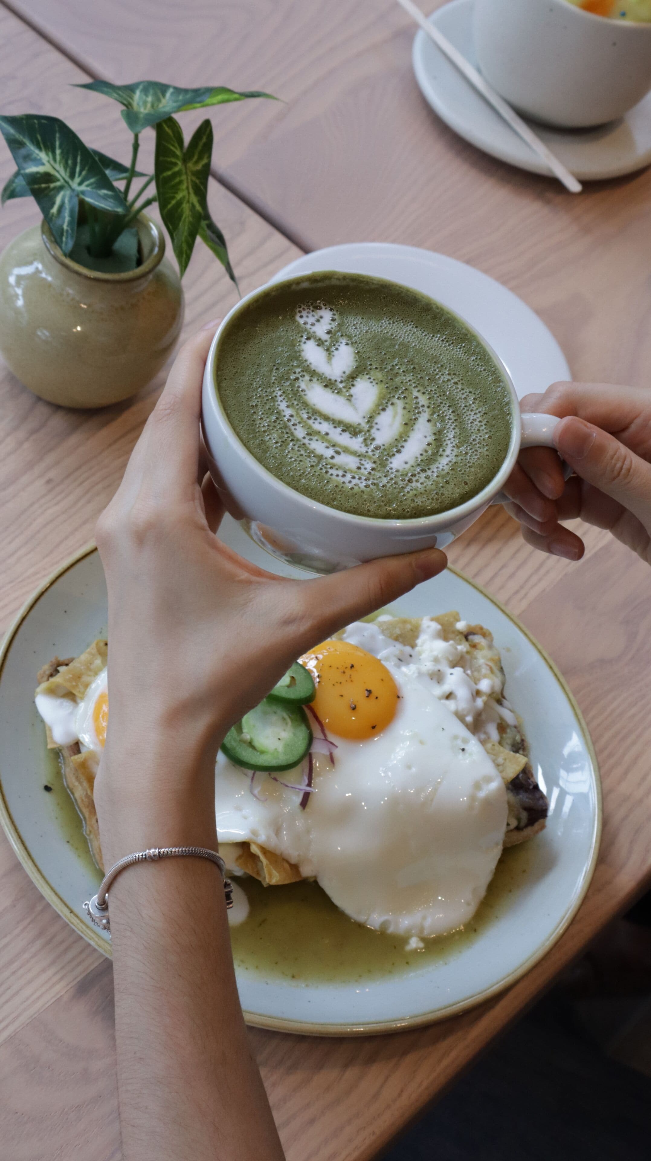 Una persona sosteniendo una taza de café verde con arte latte, un plato con huevo y aguacate, y una planta pequeña sobre la mesa.