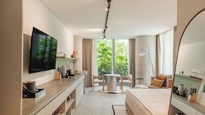 Een moderne hotelkamer met een flatscreen televisie, een ronde eettafel, een bank en een groot raam met uitzicht op groen.