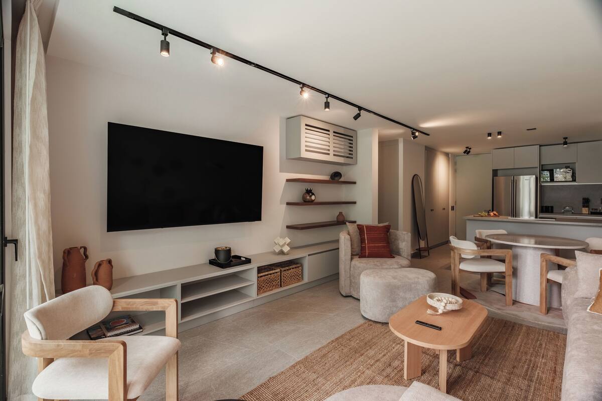 A modern living room with a flat-screen TV, wooden furniture, a kitchen area with a dining table, and built-in shelving.