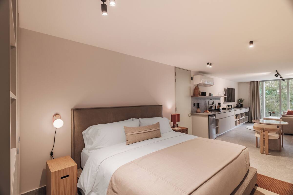 A modern hotel room with a large bed, a bedside table, a desk, a chair, and a view of the outdoors.