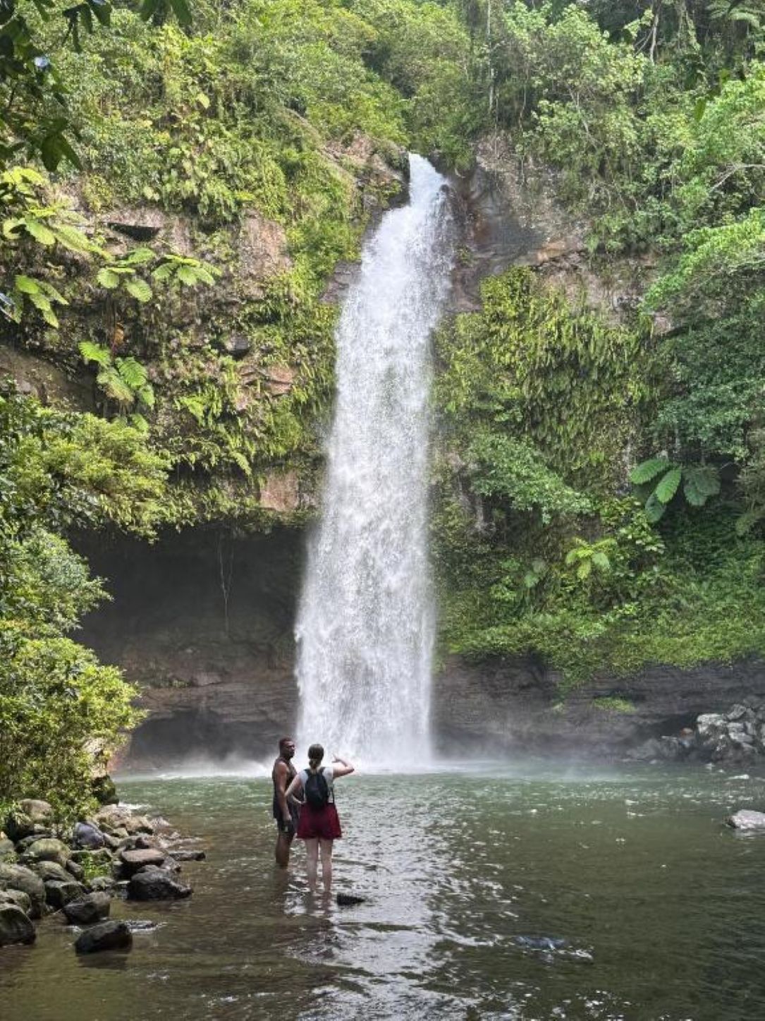 A waterfall in a forested area with two people standing in the water.