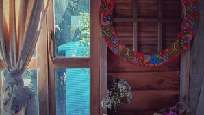 A wooden cabin interior with a window, a decorative mirror, and a small shelf with a vase and a basket. - Nisarga Pod House Kaptai (Rajasthali)