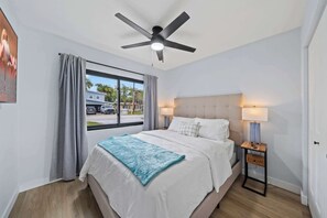 A bedroom with a bed, a ceiling fan, a nightstand, a window with curtains, and a view of the outside. - Cocoa Beach 4BR/2BA Retreat w Heated Pool (Cocoa Beach)