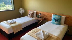 Two single beds with white linens, a wooden headboard, a bedside table with a lamp, a fan, and a window with a view of trees.