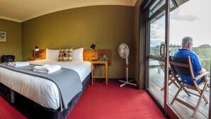 A hotel room with a large bed, a bedside table, a fan, a view of the outdoors, and a balcony with a chair. - Timber Trail Lodge & Transport (Piropiro)