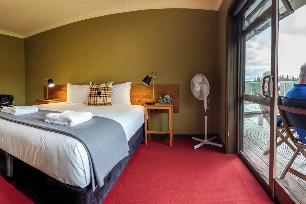 Habitación de hotel con una cama grande, una mesita de noche, un ventilador, vistas al exterior y un balcón con una silla.
