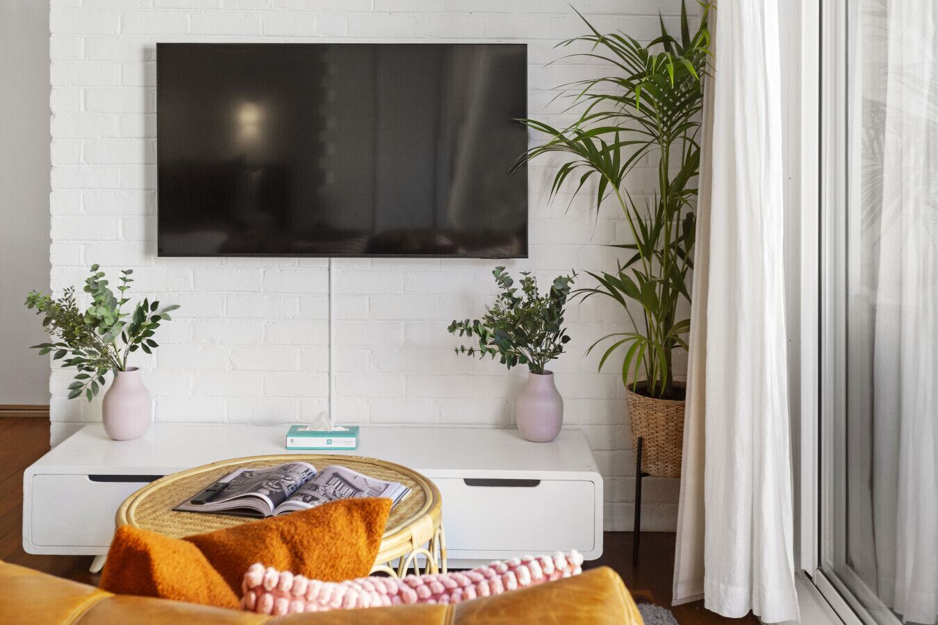 Un salón con una televisión de pantalla plana montada en una pared de ladrillo blanco, un sofá con una manta, dos plantas en macetas y una mesita auxiliar con revistas.
