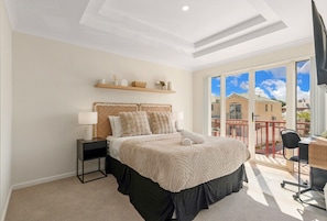 A bedroom with a bed, bedside tables, a desk, and a view of buildings through large windows.