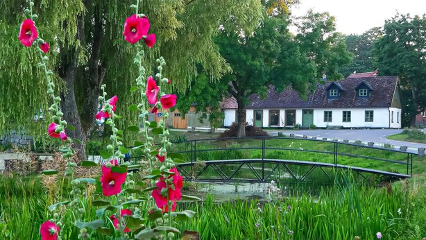 Ein Garten mit einer Brücke, einem Teich und einem weißen Gebäude mit rotem Dach.