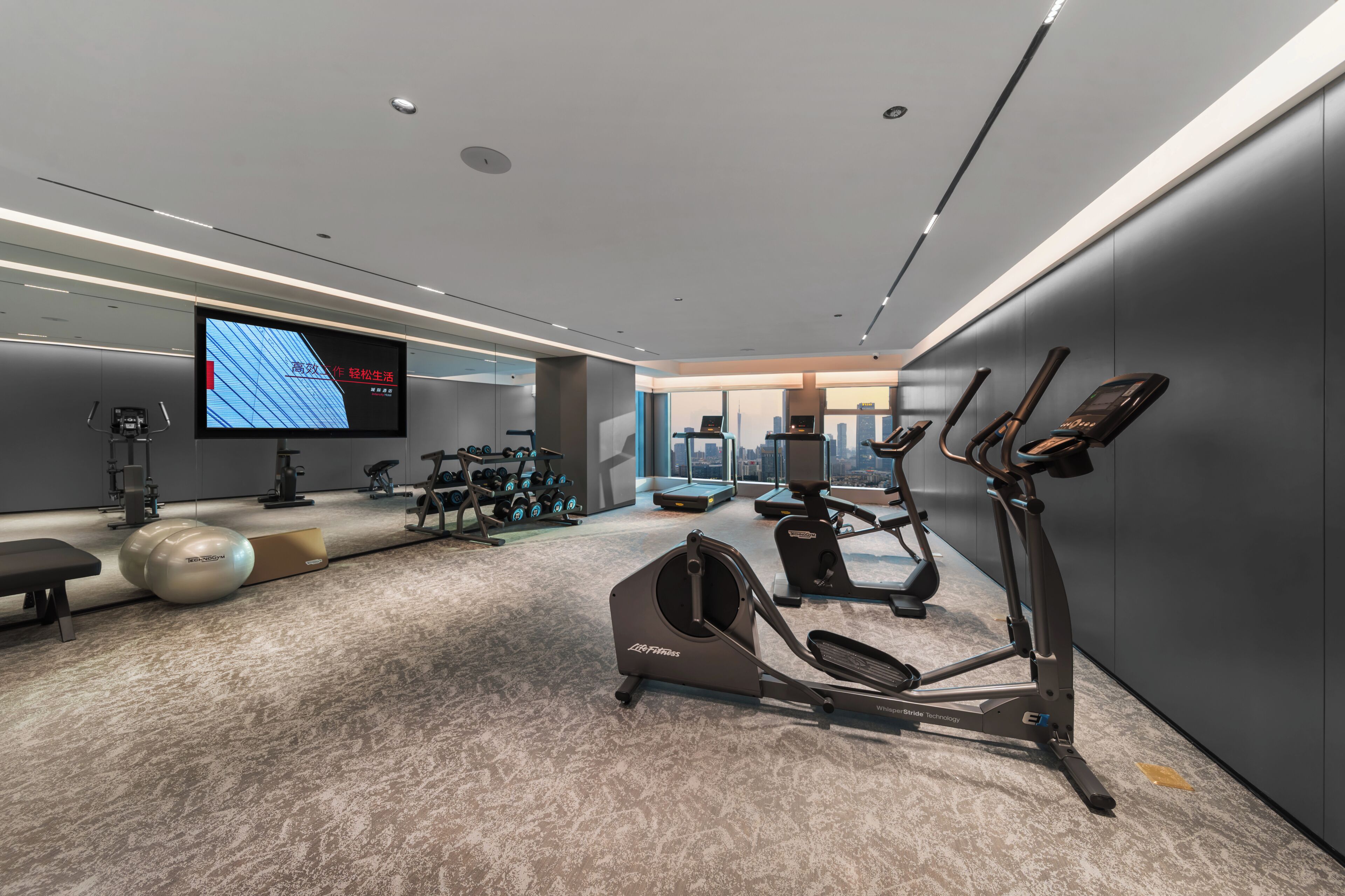 Et moderne fitnesscenter med løbebånd, styrketræningsudstyr og et stort skærm, der viser information.