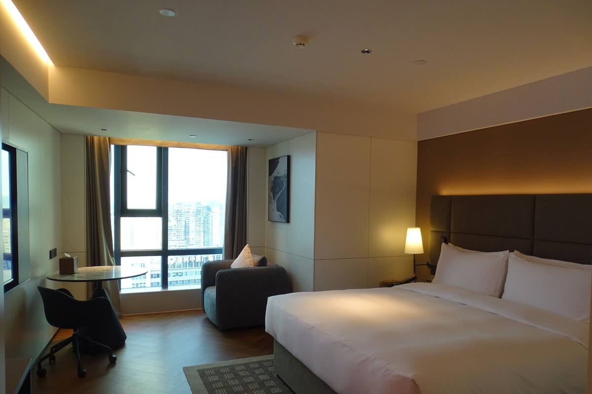Een hotelkamer met een bed, een bureau, een stoel, een lamp en een raam met uitzicht op de stad.