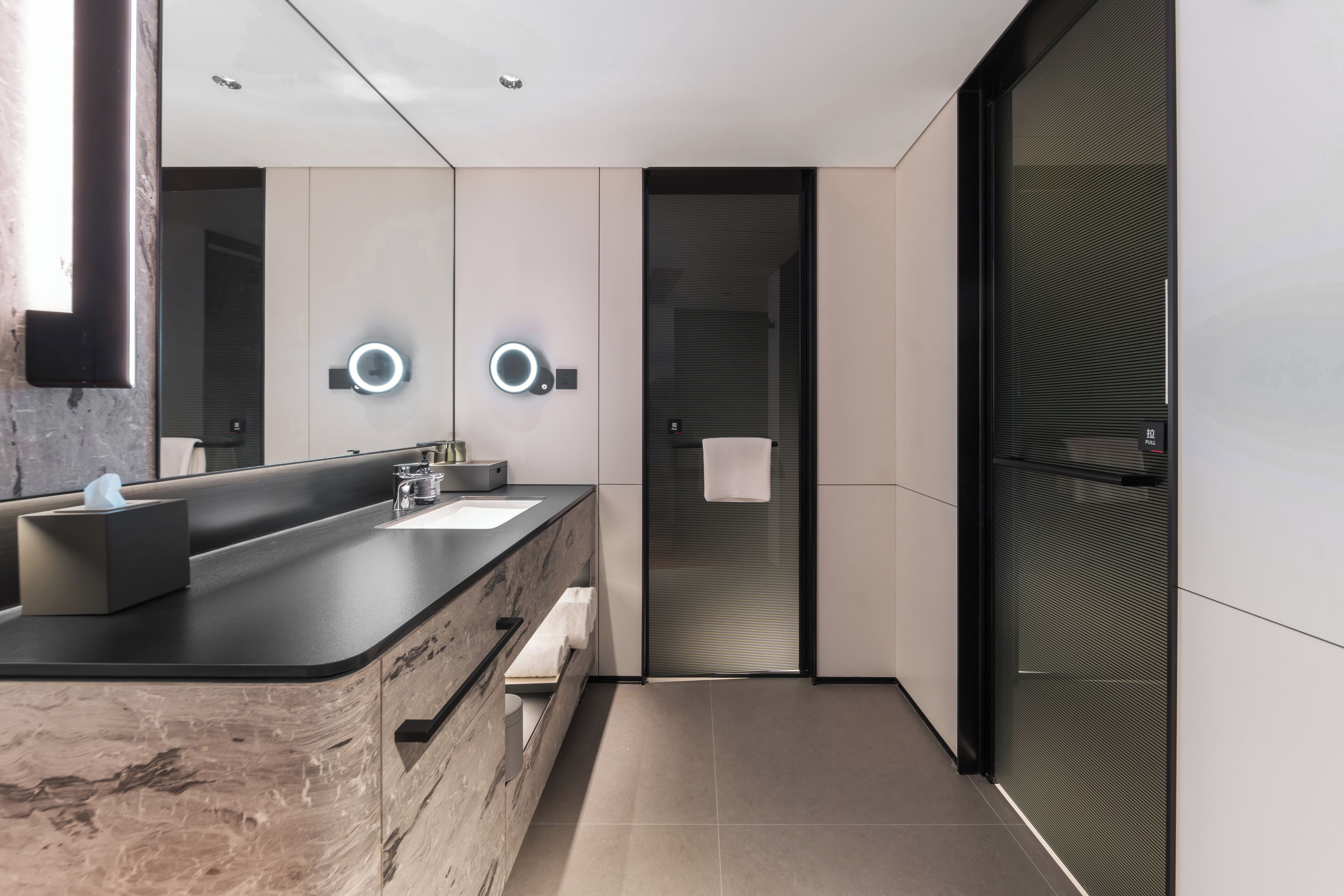 A modern bathroom with a marble vanity, two illuminated wall sconces, a glass shower enclosure, and a towel rack.