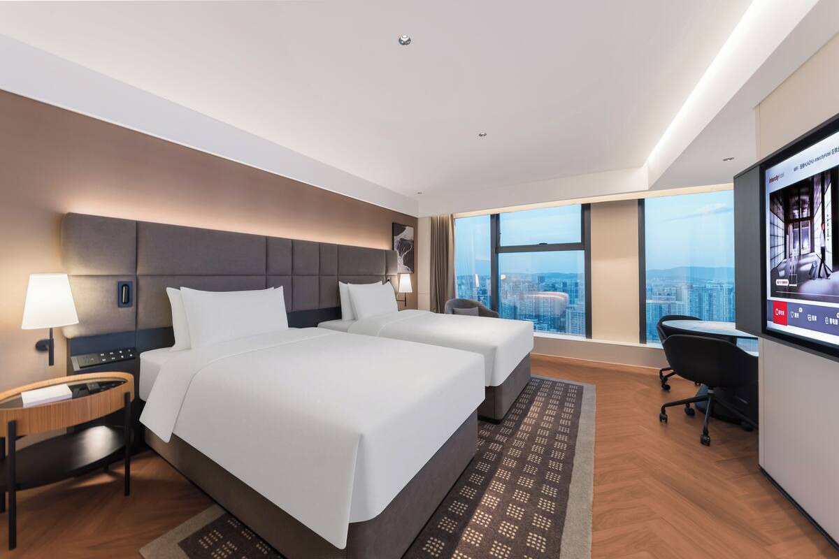 Een hotelkamer met twee bedden, een bureau, een stoel, een televisie en uitzicht over de stad.