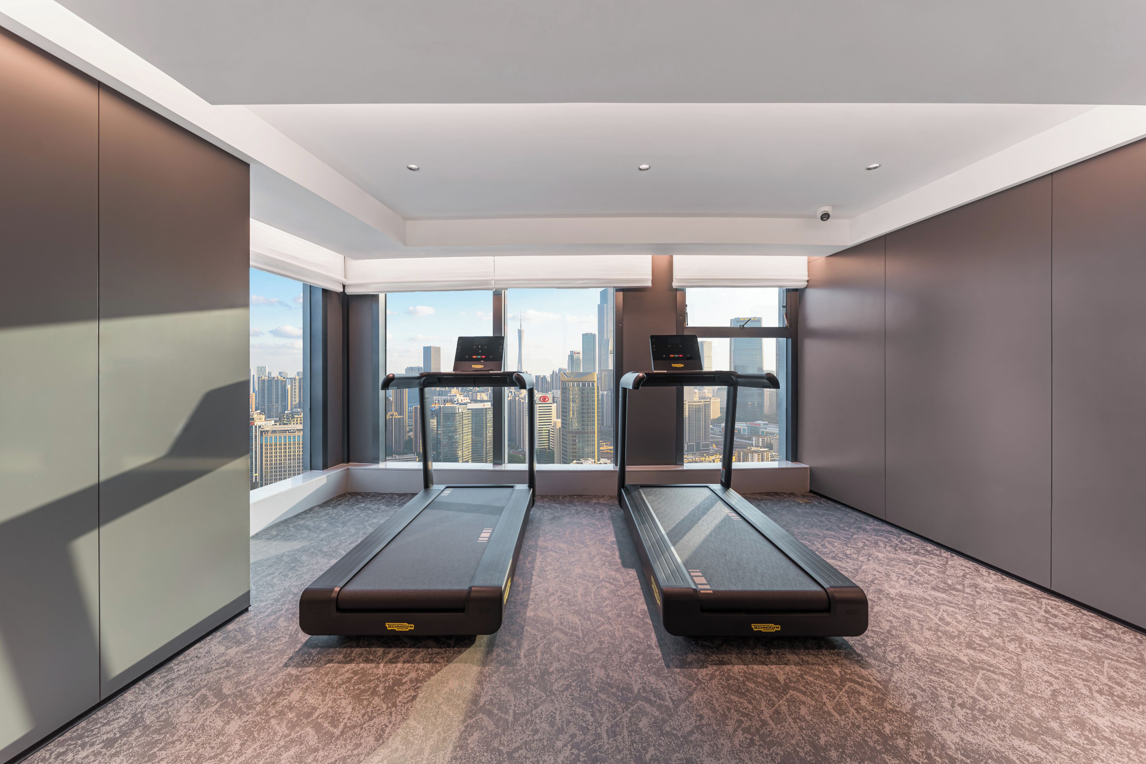 Una habitación moderna con dos cintas de correr, ventanas amplias con vista a la ciudad y un techo alto.