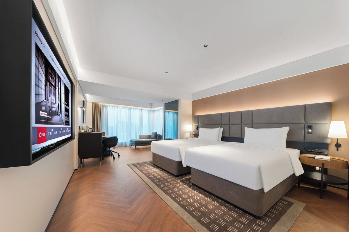 A hotel room with a large bed, a desk, a chair, a TV mounted on the wall, and a window with curtains.