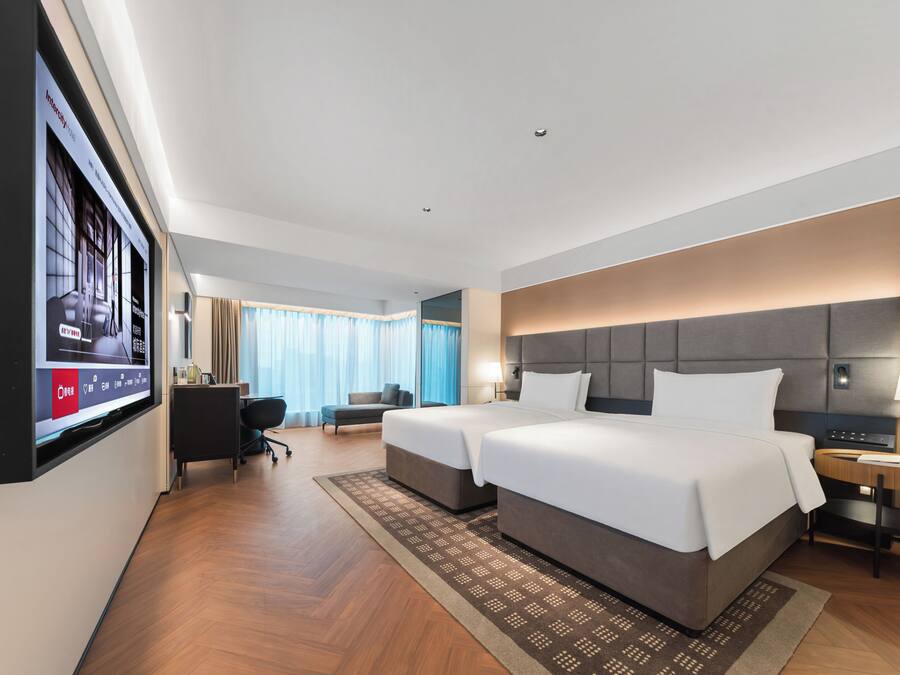 A hotel room with a large bed, a desk, a chair, a TV mounted on the wall, and a window with curtains.