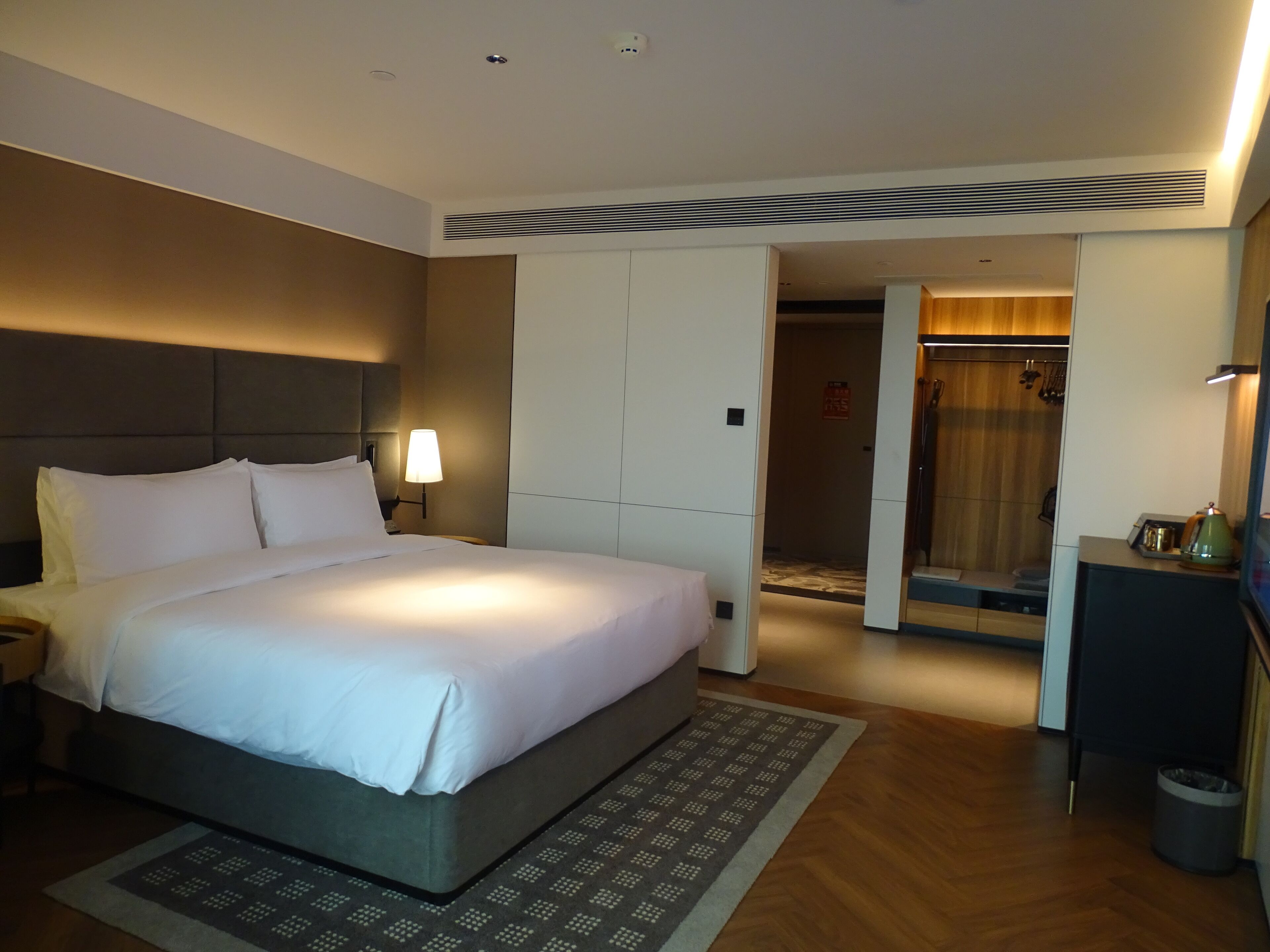 A hotel room with a large bed, a nightstand, a desk with a lamp, a wardrobe, and a shower area.