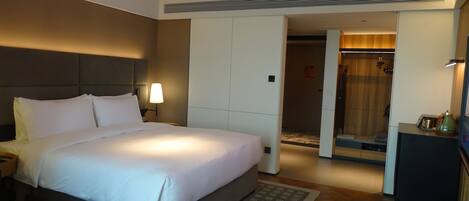 A hotel room with a large bed, a nightstand, a desk with a lamp, a wardrobe, and a shower area.