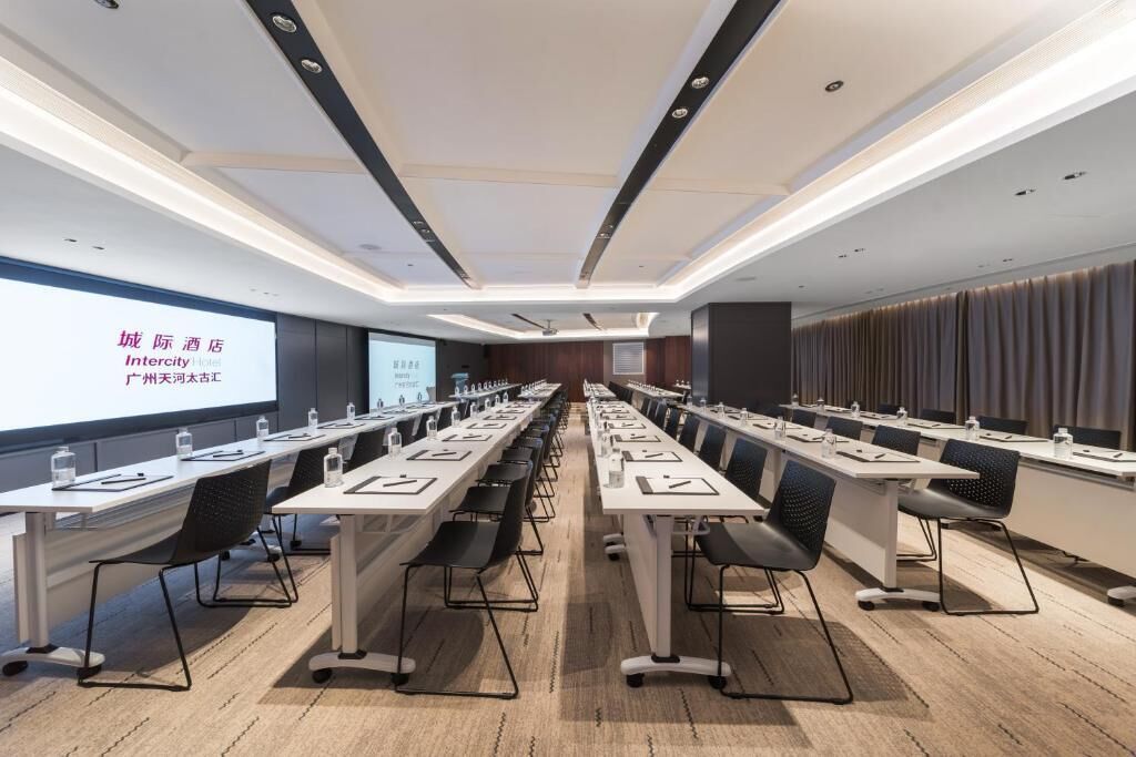 Un salón de conferencias con mesas largas, sillas y una pantalla para presentaciones.