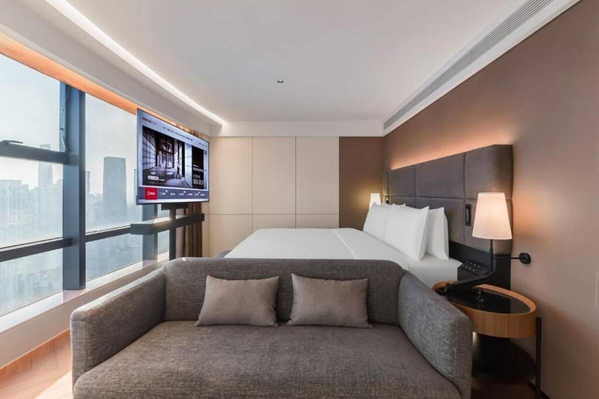 Een moderne hotelkamer met een groot bed, een bank, een aan de muur bevestigde televisie en uitzicht op de stad door grote ramen.