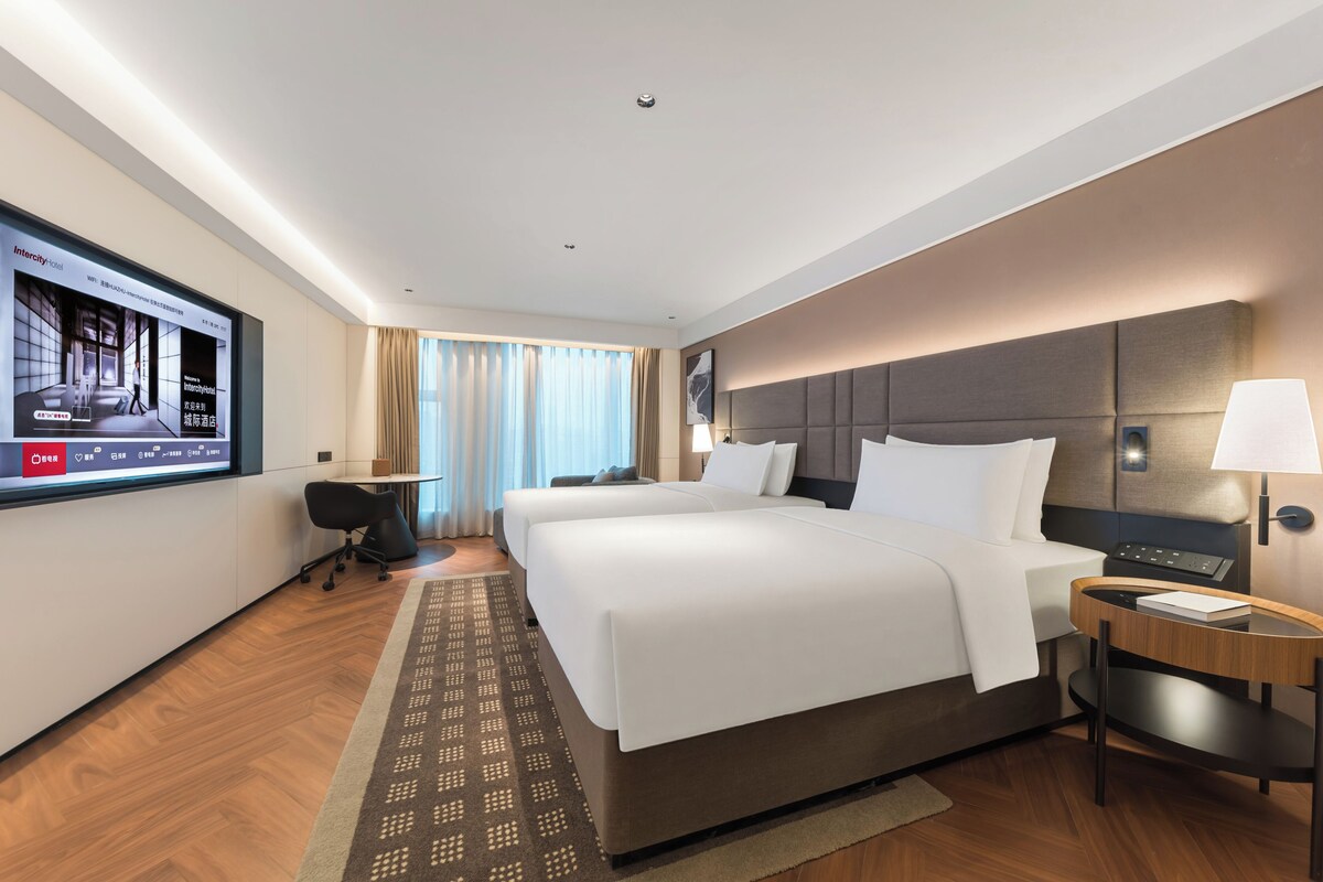 Een hotelkamer met een grote flatscreen televisie, twee bedden, een bureau met een lamp en een kamerkleed met een patroon.