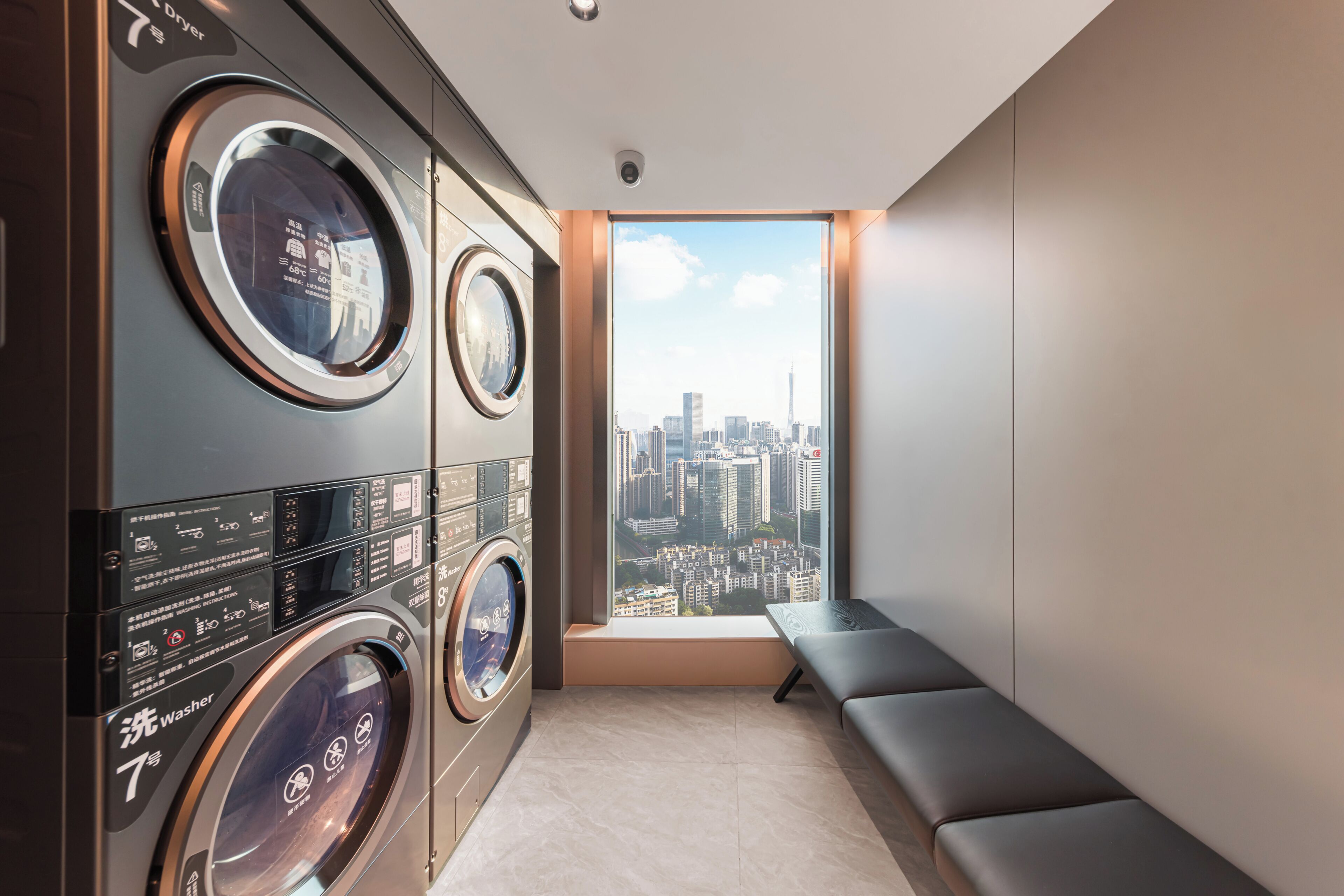 A laundry room with multiple washing machines, a bench, and a city view through a window.