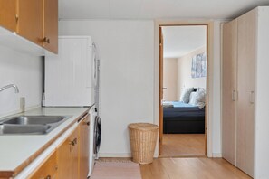 A compact kitchen with a washing machine, a small wicker basket, and a bed visible through an open door.