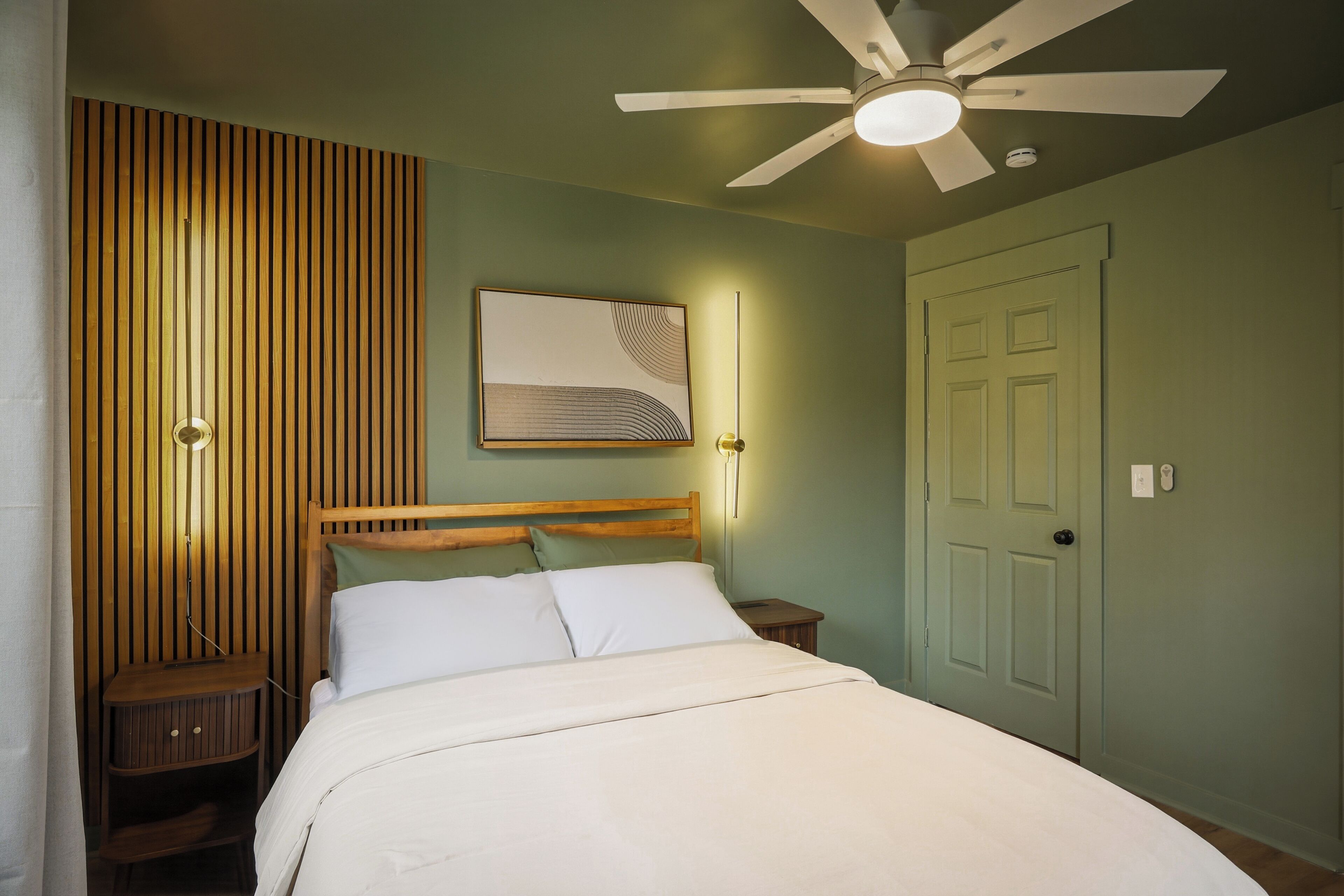 Une chambre à coucher avec un lit, un ventilateur de plafond, une lampe fixée au mur et une porte.