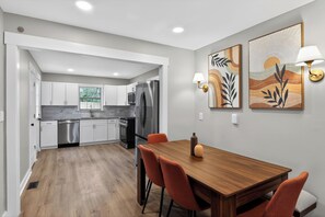 A modern kitchen with white cabinets, stainless steel appliances, and a wooden dining table with orange chairs.