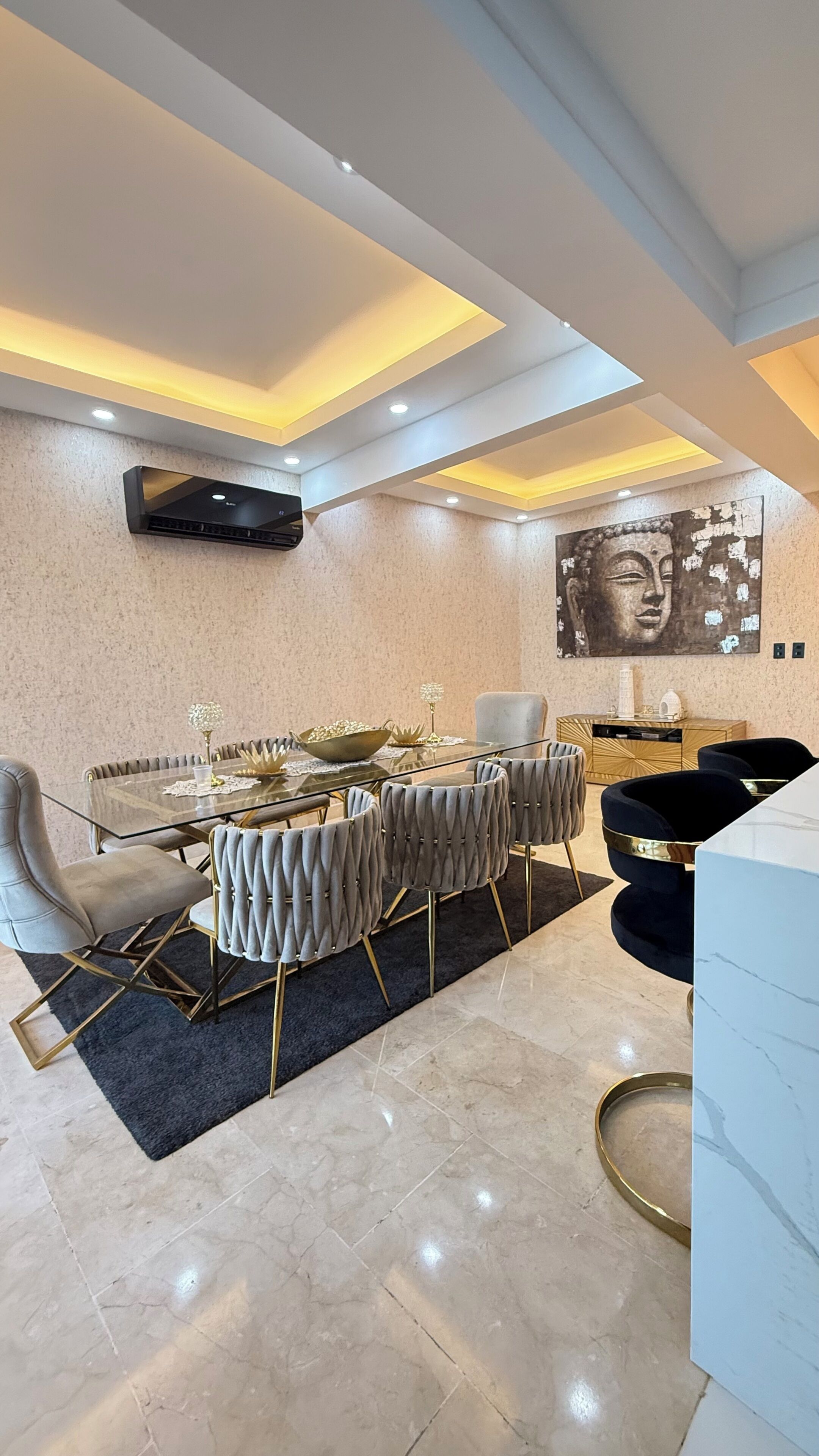 A modern dining area with a glass table, upholstered chairs, a marble floor, recessed lighting, and a wall-mounted air conditioning unit.