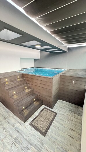 Une piscine intérieure moderne, dotée d’une terrasse en bois et de marches menant à l’eau, entourée d’un sol carrelé et d’un tapis à motifs.