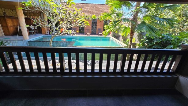 A pool with a wooden deck, surrounded by tropical plants and a building with a tiled roof. - Kami House Ubud (Ubud)
