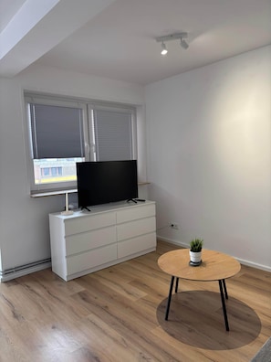 Un salon moderne avec un téléviseur à écran plat posé sur un meuble blanc, une table ronde en bois et une plante en pot.