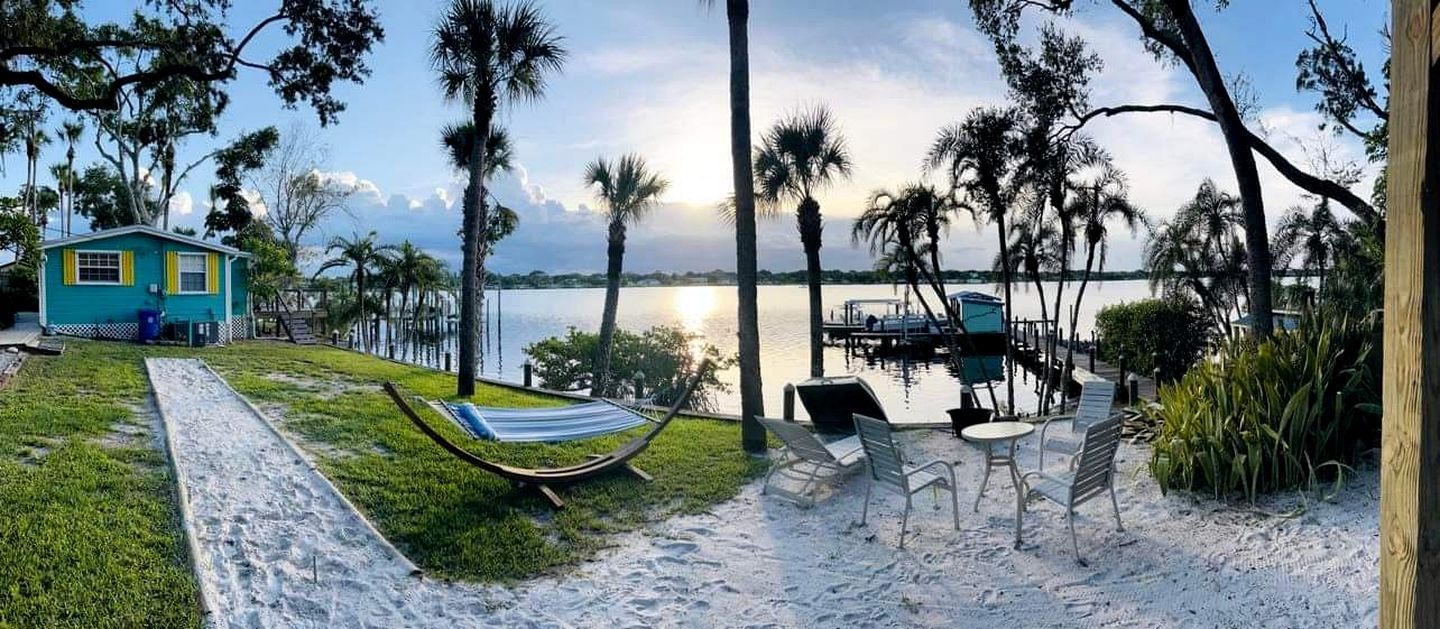 A beachside view with a hammock, chairs, and a small house by the water.