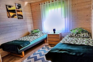 Una habitación tipo cabaña con dos camas individuales, piso de madera, un ventanal con cortinas traslúcidas y dos cuadros colgados en la pared.
