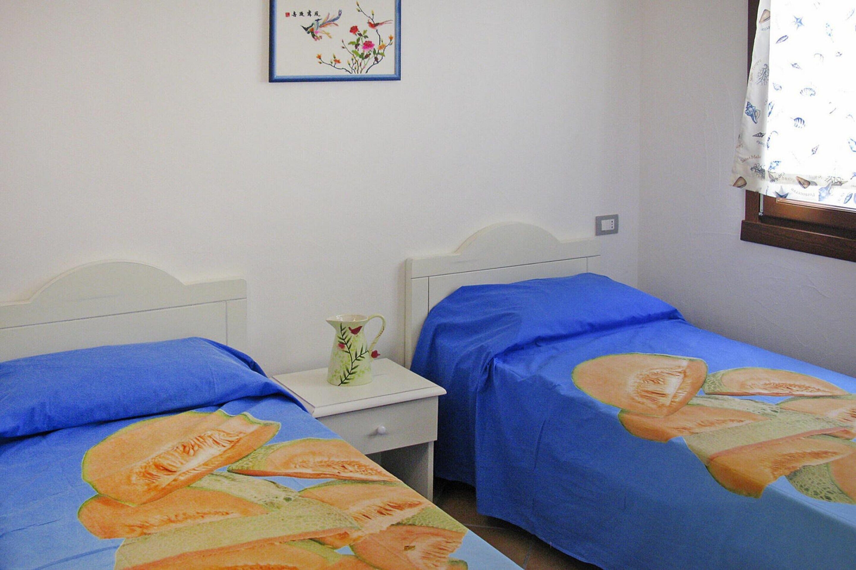 A bedroom with two single beds, each with a blue bedspread featuring a fruit design. There is a white nightstand with a pitcher and a framed picture on the wall above the beds.