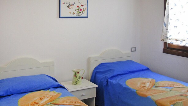 Een slaapkamer met twee eenpersoonsbedden, elk met een blauwe bedspreide met een fruitmotief. Er staat een witte nachtkastje met een kan en er hangt een lijst aan de muur boven de bedden.