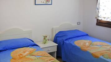 A bedroom with two single beds, each with a blue bedspread featuring a fruit design. There is a white nightstand with a pitcher and a framed picture on the wall above the beds.