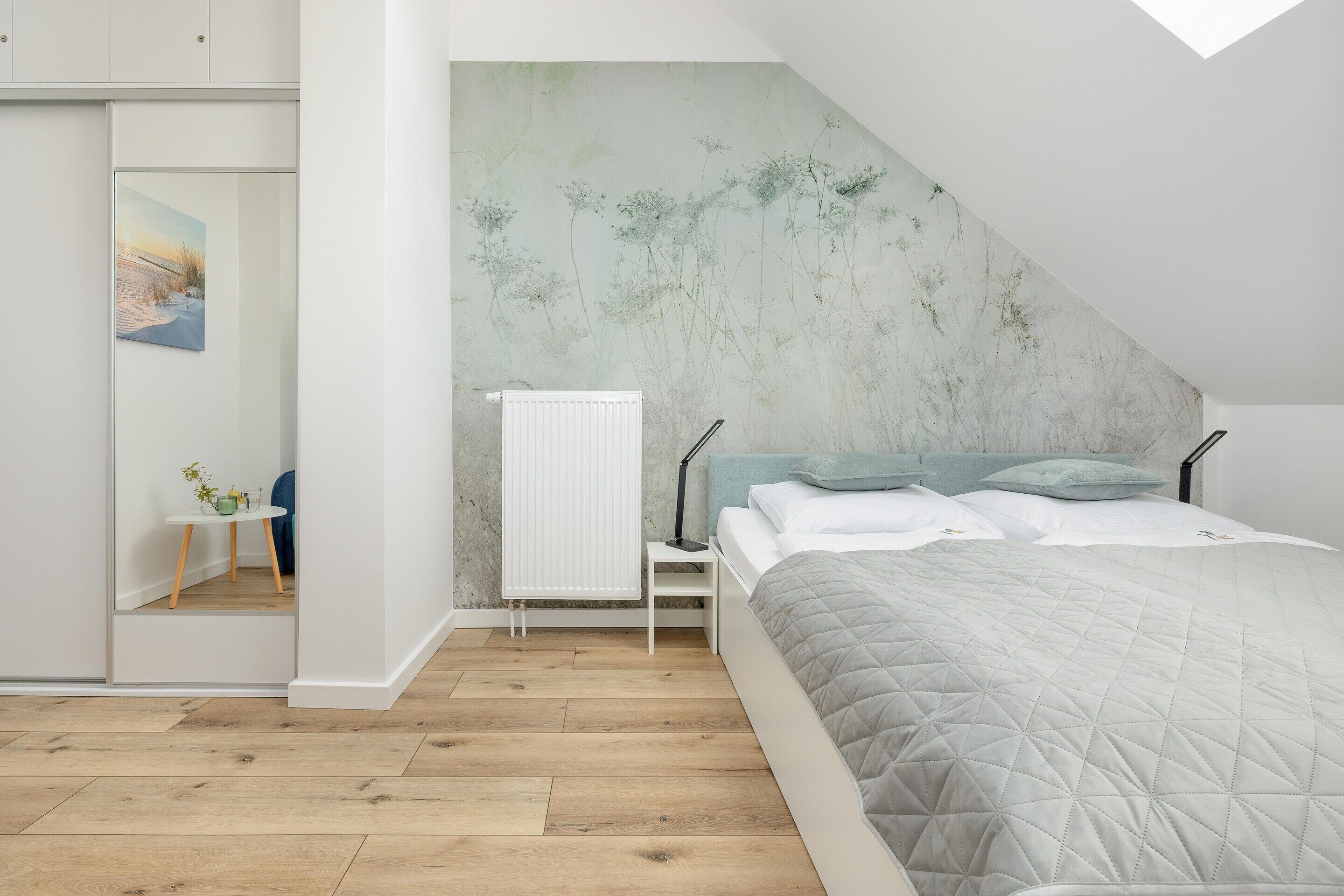 A bedroom with a bed, a radiator, a small table with a plant, and a painting on the wall.