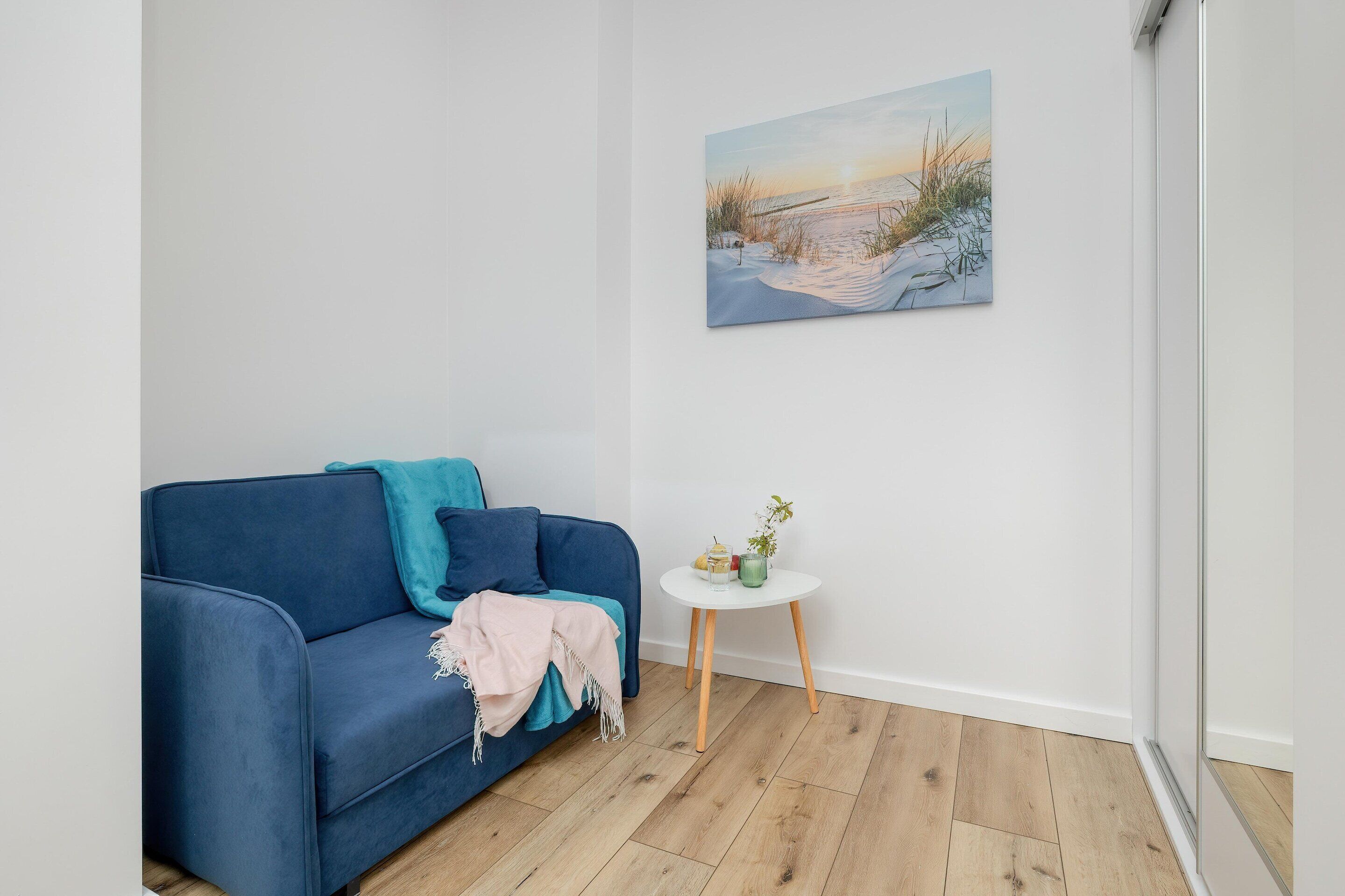Quarto com um sofá azul, uma mesinha branca com uma planta e um quadro de uma praia na parede.